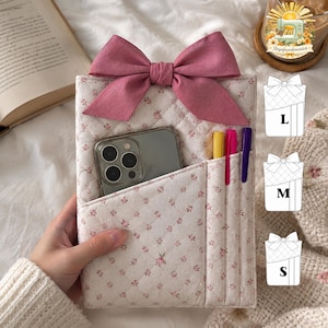 Può includere: Una custodia organizer in tessuto trapuntato con un piccolo motivo floreale rosa su sfondo bianco, sormontata da un grande fiocco in tessuto rosa. Include una tasca frontale con uno smartphone e tre scomparti verticali con penne gialla, rosa e viola.