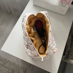 Pu&ograve; includere: Una pochette in tessuto trapuntato con esterno bianco e un delicato motivo floreale rosa e arancione. L'interno &egrave; foderato con un tessuto a quadretti vichy giallo e bianco. Una spazzola elettrica nera &egrave; riposta all'interno della pochette.