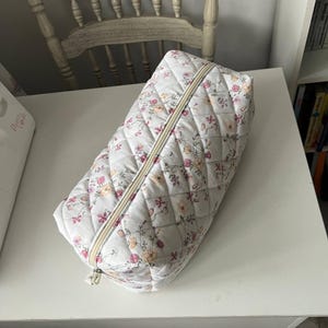 Pu&ograve; includere: Una pochette trapuntata rettangolare con base bianca e un delicato motivo floreale nei toni del rosa, arancione e verde. La borsa presenta una texture con cuciture a rombi e una cerniera centrale color crema lungo la parte superiore.