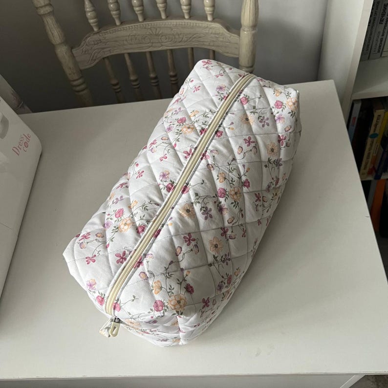 Pu&ograve; includere: Una pochette trapuntata rettangolare con base bianca e un delicato motivo floreale nei toni del rosa, arancione e verde. La borsa presenta una texture con cuciture a rombi e una cerniera centrale color crema lungo la parte superiore.
