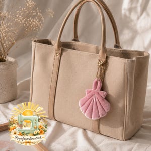 May include: Beige canvas tote bag with tan leather handles and accents. A pink velvet seashell charm hangs from the handle. The bag features the text "Rayofsunshinestitch" on a logo with a sewing machine.
