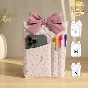 May include: A quilted fabric pouch featuring a light pink floral pattern and a large mauve bow at the top. The pouch has a front pocket containing a smartphone and three pens in yellow, pink, and purple. A size chart shows L, M, and S options.