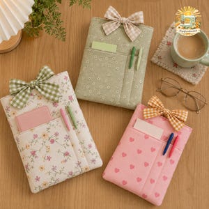 May include: Three fabric notebook covers featuring floral and heart patterns, each adorned with a gingham bow. The covers include front pockets containing small cards and pens. The designs include a sage green floral, a cream floral, and a pink heart print.