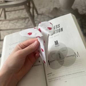 Può includere: Un segnalibro in tessuto bianco con un motivo ripetuto di piccoli cuori rossi. Il segnalibro è piegato a forma di V ed è posizionato tra le pagine di un libro aperto che mostra del testo e un diagramma circolare con la parola Limitless.