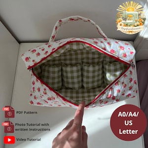 Puede incluir: Un bolso de tela acolchada con base blanca, estampado floral rojo y un asa superior. El interior está forrado con un patrón de cuadros vichy verde y blanco e incluye cuatro bolsillos internos. Una cremallera roja recorre la abertura superior.