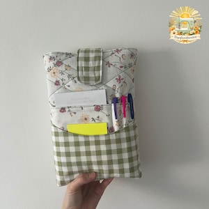 May include: A quilted fabric pouch featuring a white floral pattern with green gingham accents. The pouch includes a top flap closure, a front pocket containing a white card and a bright yellow sticky note, and three pens in blue, pink, and purple.