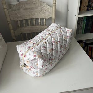 Pu&ograve; includere: Una trousse trapuntata rettangolare con base in tessuto bianco e un delicato motivo floreale nei toni del rosa, giallo e verde. La borsa presenta una texture con cuciture a rombi e una chiusura a cerniera beige lungo il bordo superiore.