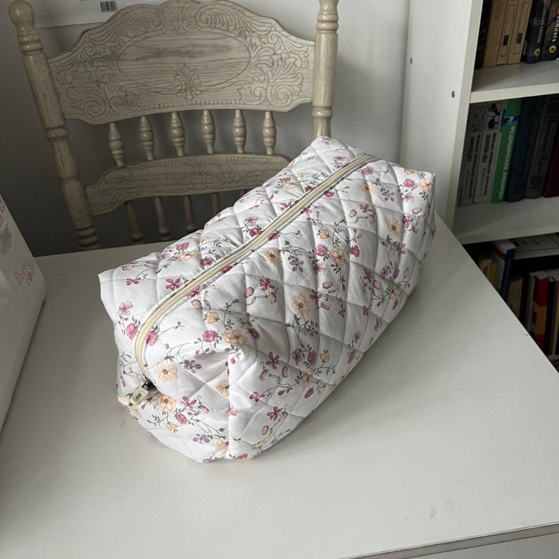 Pu&ograve; includere: Una trousse trapuntata rettangolare con base in tessuto bianco e un delicato motivo floreale nei toni del rosa, giallo e verde. La borsa presenta una texture con cuciture a rombi e una chiusura a cerniera beige lungo il bordo superiore.