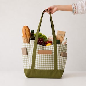 May include: A sage green and white gingham tote bag with a solid olive green base and long shoulder straps. The bag features three exterior pockets containing a small notebook and two small cases, and is filled with groceries including bread.