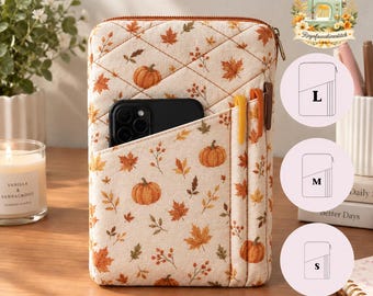 Puede incluir: Una funda de tela acolchada para tableta con fondo crema y un patrón otoñal de calabazas naranjas, hojas de arce marrones y bayas. La funda incluye un bolsillo frontal que contiene un teléfono inteligente negro y dos ranuras para bolígrafos.