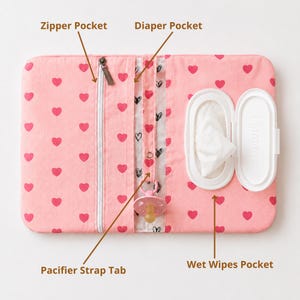 Puede incluir: Un organizador rectangular de tela rosa con un patrón repetitivo de corazones rojos. Incluye un bolsillo con cremallera, un bolsillo para pañales, una lengüeta para chupete con un chupete rosa y un bolsillo para toallitas con tapa abierta.