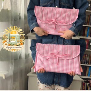 Puede incluir: Una funda rectangular de tela acolchada con un patrón de cuadros vichy en rosa y blanco. La funda tiene un cierre de solapa redondeado asegurado con un lazo de tela atado en el centro. El exterior está hecho de material suave y acolchado con costuras verticales.