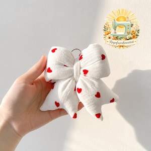 May include: A white fabric bow keychain featuring a repeating pattern of small red hearts. The bow is constructed with a soft, textured material and includes a silver metal ring at the top for attachment. The design is simple and symmetrical.