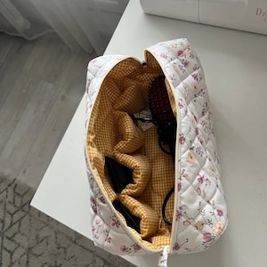 Pu&ograve; includere: Una borsa da toilette rettangolare trapuntata con esterno bianco e un delicato motivo floreale nei toni del rosa, arancione e verde. L'interno &egrave; foderato con un tessuto a quadretti giallo e bianco e include diverse tasche interne e una cerniera.
