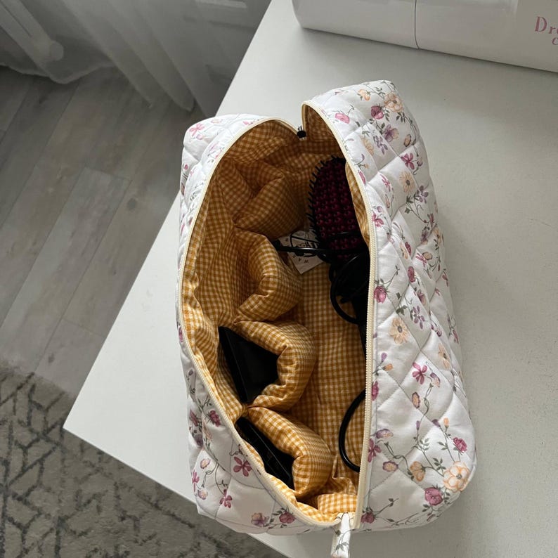 Pu&ograve; includere: Una borsa da toilette rettangolare trapuntata con esterno bianco e un delicato motivo floreale nei toni del rosa, arancione e verde. L'interno &egrave; foderato con un tessuto a quadretti giallo e bianco e include diverse tasche interne e una cerniera.