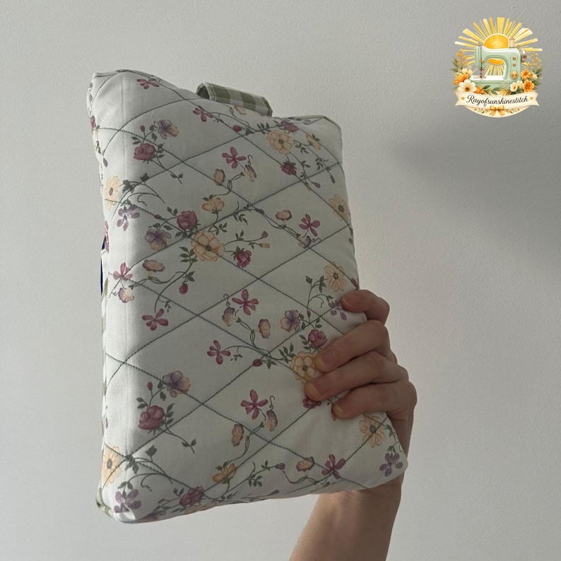 May include: A rectangular quilted fabric pouch featuring a white base with a delicate floral pattern in shades of pink, yellow, and green. The fabric is stitched in a diamond grid pattern and includes a small green and white checkered fabric tab.