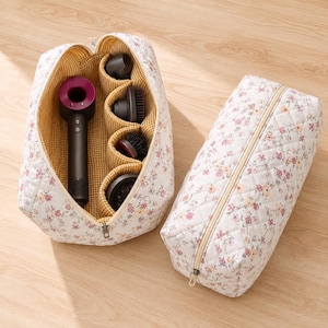 Puede incluir: Dos bolsas de almacenamiento acolchadas con estampado floral blanco y cremalleras beige. Una bolsa está abierta, revelando un interior a cuadros amarillos y blancos que contiene un secador de pelo gris oscuro y cuatro accesorios negros.