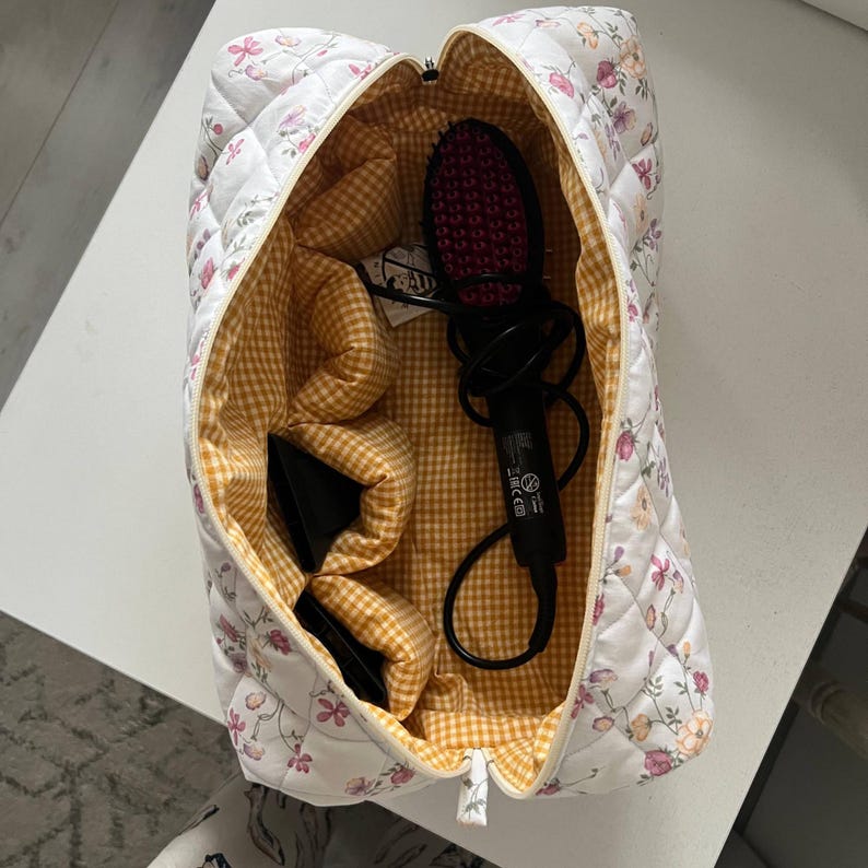 Pu&ograve; includere: Una borsa da viaggio trapuntata bianca con motivo floreale e fodera interna a quadretti vichy gialli e bianchi. La borsa aperta contiene uno strumento per lo styling dei capelli nero con setole rosa e due beccucci neri per asciugacapelli.