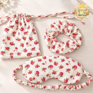 May include: A white fabric set featuring a repeating pattern of small red roses and green leaves. The set includes a drawstring pouch, a circular hair scrunchie, and a contoured sleep mask with an elastic strap, all made from the same floral textile.
