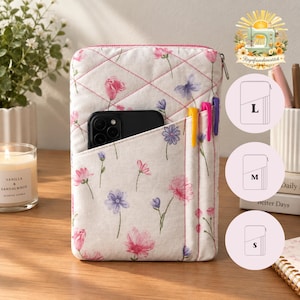 May include: A quilted fabric pouch featuring a white background with a pink and purple floral pattern. The pouch has a front pocket containing a black smartphone and two pens, with a zip closure at the top. Three circular size icons labelled L, M, and S are visible.