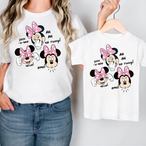 Op de afbeelding: Witte T-shirts met een Minnie Mouse-afbeelding. De afbeelding toont Minnie Mouse met een roze strik en de zinnen "Peek-a-boo", "Hello!", "OMG!" en "Ah, ah, so funny!" op de shirts.