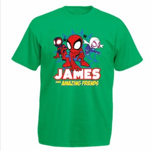 May include: A bright green short-sleeve t-shirt featuring a graphic of three cartoon superheroes in front of a blue spiderweb. Below the characters, the text reads JAMES AND AMAZING FRIENDS in bold white and yellow lettering on the front.