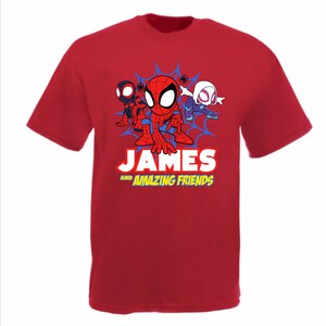 May include: A red short-sleeved t-shirt featuring a graphic of three cartoon superheroes in front of a blue spiderweb. Below the characters, the text reads JAMES AND AMAZING FRIENDS in white and yellow block lettering on the front of the shirt.