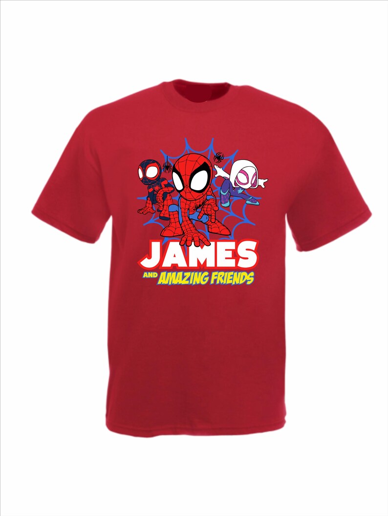 May include: A red short-sleeved t-shirt featuring a graphic of three cartoon superheroes in front of a blue spiderweb. Below the characters, the text reads JAMES AND AMAZING FRIENDS in white and yellow block lettering on the front of the shirt.