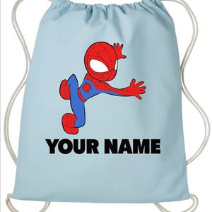 May include: A light blue drawstring backpack featuring a cartoon illustration of a red and blue superhero character in a dynamic pose. Below the character, the text YOUR NAME is printed in bold black capital letters. The bag has white cords.