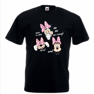 Op de afbeelding: Zwart T-shirt met een Minnie Mouse-ontwerp. Het ontwerp toont Minnie in drie verschillende poses, elk met een roze strik en tekstballonnen met de tekst "Peek-a-boo", "Hello!", "Ah, ah, so funny!" en "omg!".