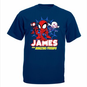 May include: A dark blue short-sleeved t-shirt featuring a graphic of three cartoon superheroes in front of a blue spiderweb. Below the characters, the text reads JAMES AND AMAZING FRIENDS in bold white and yellow lettering with a red outline.