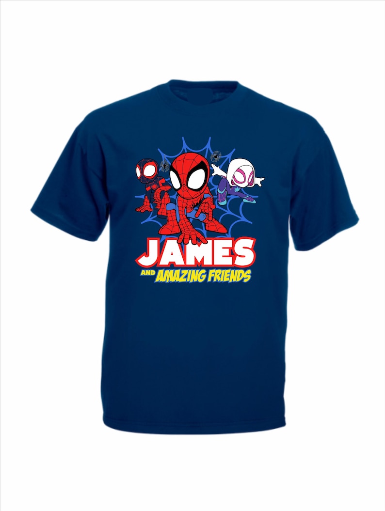 May include: A dark blue short-sleeved t-shirt featuring a graphic of three cartoon superheroes in front of a blue spiderweb. Below the characters, the text reads JAMES AND AMAZING FRIENDS in bold white and yellow lettering with a red outline.