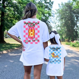 Peut inclure: Deux t-shirts blancs avec des imprimés graphiques. Le plus grand présente un motif à damier rose et bleu clair, une illustration de bouteille de sauce piquante et le texte "MAMACITA CLUB Stay Spicy". Le plus petit a un motif à damier bleu clair, une illustration de teckel et le texte "COOL KIDS CLUB Stay Curious".