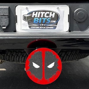 Pode incluir: Uma cobertura de engate Deadpool preta e vermelha. A cobertura apresenta um design circular com a máscara Deadpool em preto e branco sobre um fundo vermelho. A cobertura do engate está presa a um veículo preto.