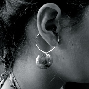 May include: A silver hoop earring with a hammered, circular pendant. The earring is worn on the ear of a person, with their hair and neck visible in the black and white image. The pendant has a textured surface.