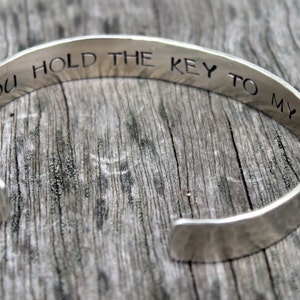 May include: Silver cuff bracelet with the engraved phrase "YOU HOLD THE KEY TO MY HEART". The open-ended bracelet is displayed on a weathered wooden surface. A simple, elegant piece of jewelry.