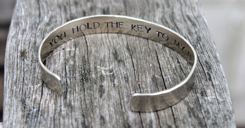 May include: Silver cuff bracelet with the engraved phrase "YOU HOLD THE KEY TO MY HEART". The open-ended bracelet is displayed on a weathered wooden surface. A simple, elegant piece of jewelry.