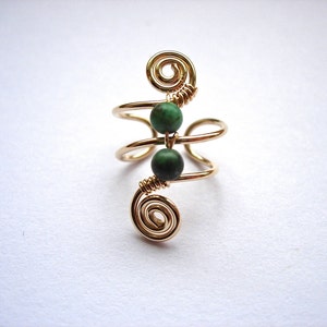 May include: A gold-colored wire ring with two dark green beads. The ring features a spiral design at the top and bottom, with the beads centered between the spirals. The ring is set against a white background.