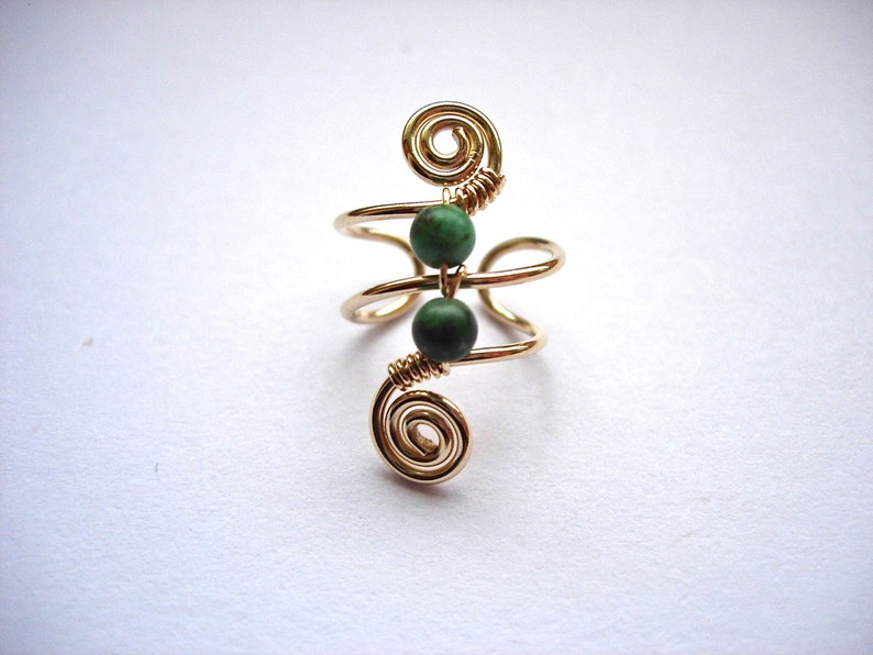 May include: A gold-colored wire ring with two dark green beads. The ring features a spiral design at the top and bottom, with the beads centered between the spirals. The ring is set against a white background.