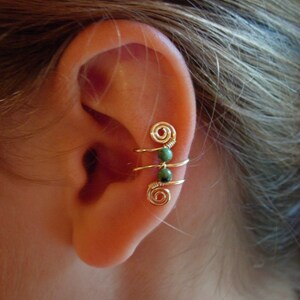 May include: Gold-colored ear cuff with a spiral design and three green beads. The ear cuff wraps around the ear, creating a unique and stylish accessory. The jewelry is shown on a person's ear.