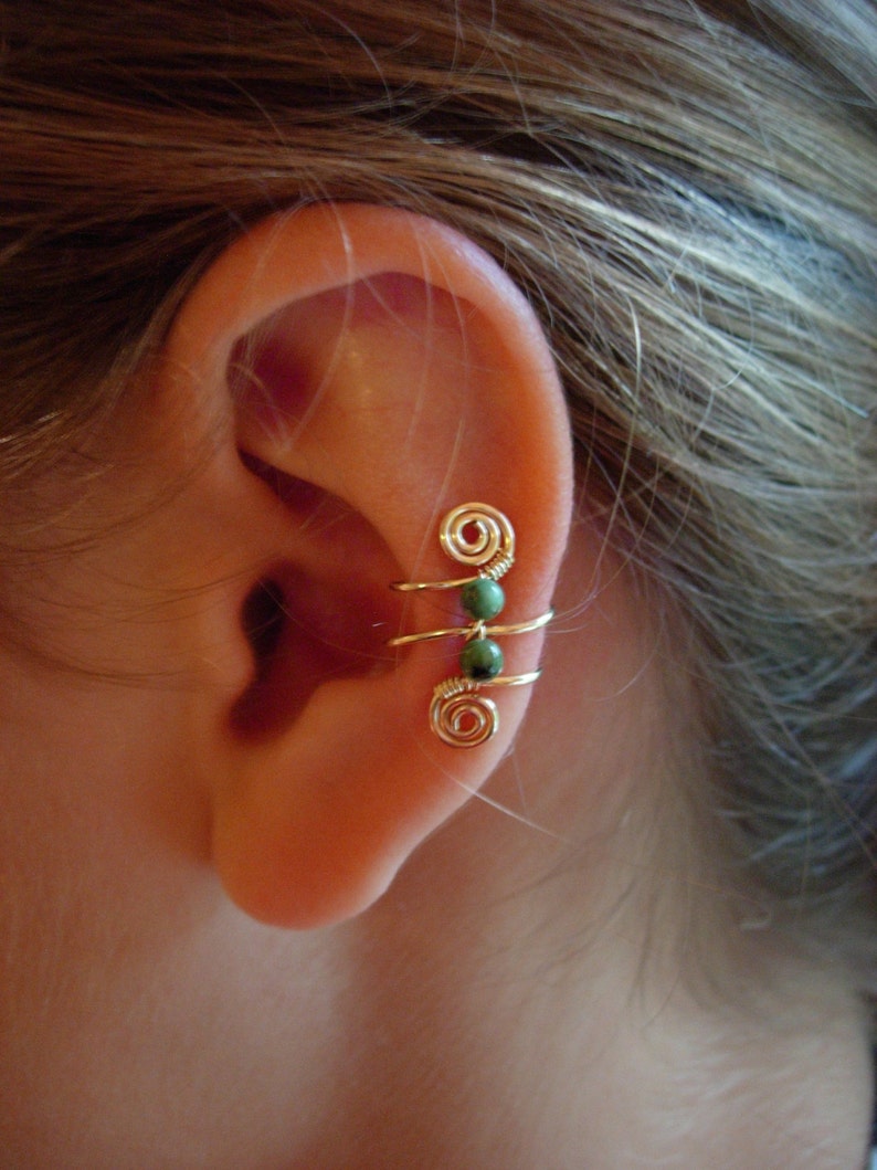 May include: Gold-colored ear cuff with a spiral design and three green beads. The ear cuff wraps around the ear, creating a unique and stylish accessory. The jewelry is shown on a person's ear.