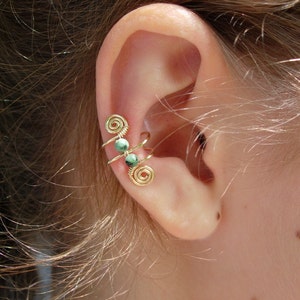 May include: A gold-colored ear cuff with a spiral design and three small green beads. The ear cuff is worn on the upper ear and is visible against the skin and hair.