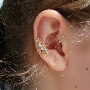 May include: Gold-colored ear cuff with a spiral design and three small, round, green beads. The ear cuff wraps around the ear, creating a unique and stylish accessory. The jewelry is shown on a person's ear.