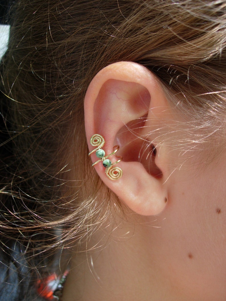 May include: Gold-colored ear cuff with a spiral design and three small, round, green beads. The ear cuff wraps around the ear, creating a unique and stylish accessory. The jewelry is shown on a person's ear.