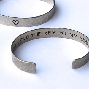 May include: Two silver-toned metal bracelets. One bracelet has a hammered texture and a small heart symbol. The other bracelet has the phrase "YOU HOLD THE KEY TO MY HEART" engraved on it. The bracelets are open cuff style.