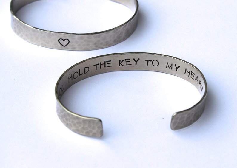 May include: Two silver-toned metal bracelets. One bracelet has a hammered texture and a small heart symbol. The other bracelet has the phrase "YOU HOLD THE KEY TO MY HEART" engraved on it. The bracelets are open cuff style.