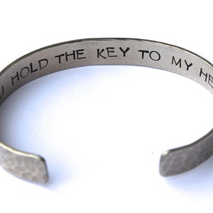 May include: Silver-toned cuff bracelet with the inscription "YOU HOLD THE KEY TO MY HEART" in a simple, sans-serif font. The bracelet has a hammered texture and an open design.
