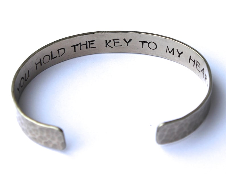 May include: Silver-toned cuff bracelet with the inscription "YOU HOLD THE KEY TO MY HEART" in a simple, sans-serif font. The bracelet has a hammered texture and an open design.