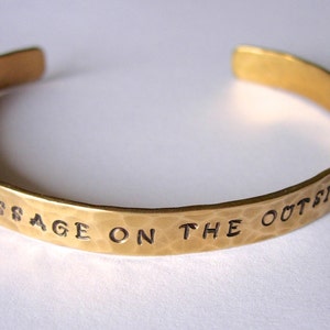 May include: A gold-colored cuff bracelet with the phrase "MESSAGE ON THE OUTSIDE" engraved in black lettering. The bracelet has a hammered texture and an open design.