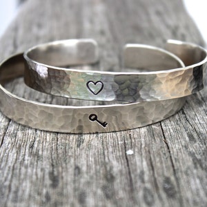 May include: Two silver-colored hammered metal cuff bracelets. One bracelet has a heart symbol, and the other has a key symbol. The bracelets are displayed on a weathered wooden surface.